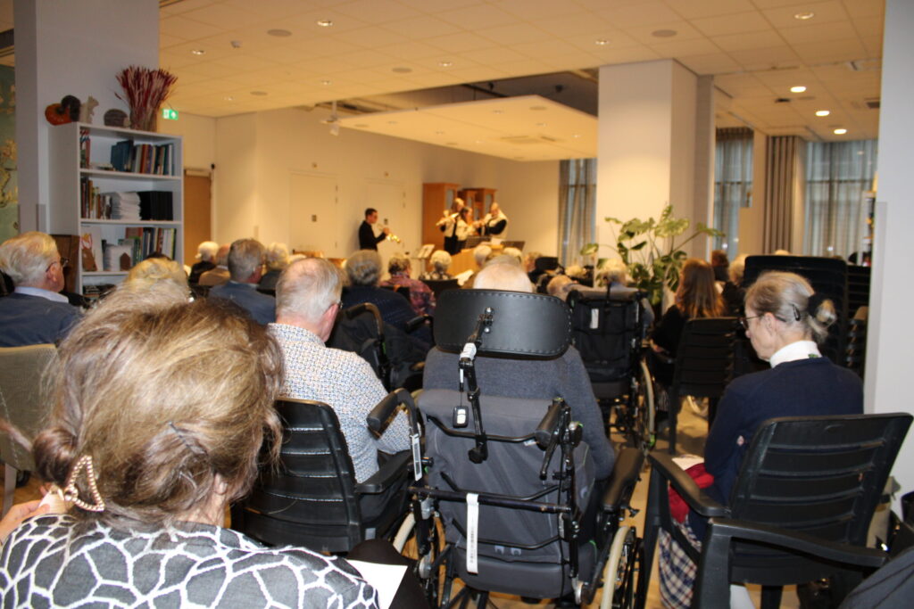 Een groep mensen zit in een zaal, kijkend naar een optreden op een podium. Er zijn rolstoelen en stoelen zichtbaar.