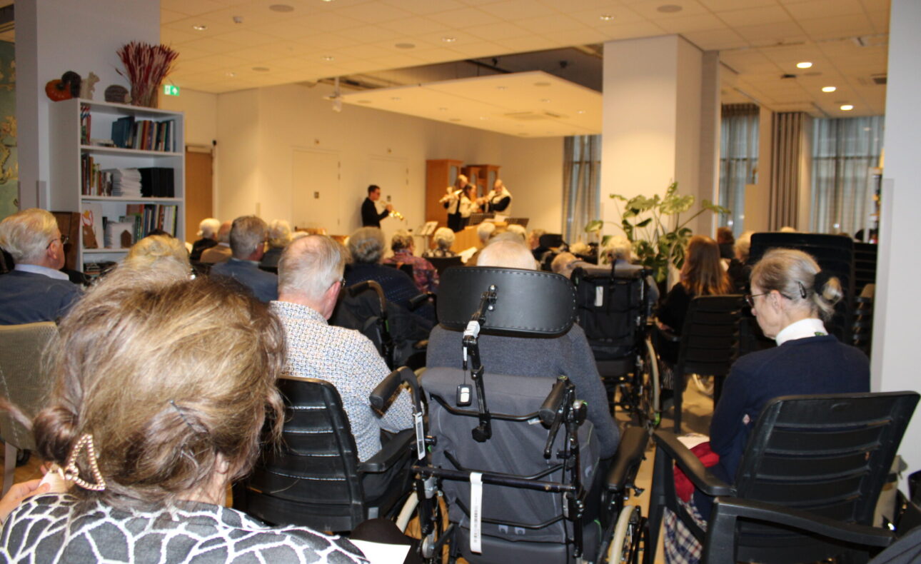 Een groep mensen zit in een zaal, kijkend naar een optreden op een podium. Er zijn rolstoelen en stoelen zichtbaar.