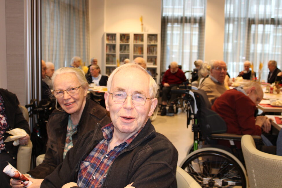 Een groep ouderen zit gezellig samen in een lichte ruimte. Ze lachen en praten met elkaar, sommigen in rolstoelen.