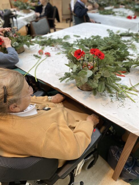 Een oudere vrouw in een rolstoel maakt een bloemstuk met rode bloemen en groen blad op een tafel in een gemeenschappelijke ruimte.
