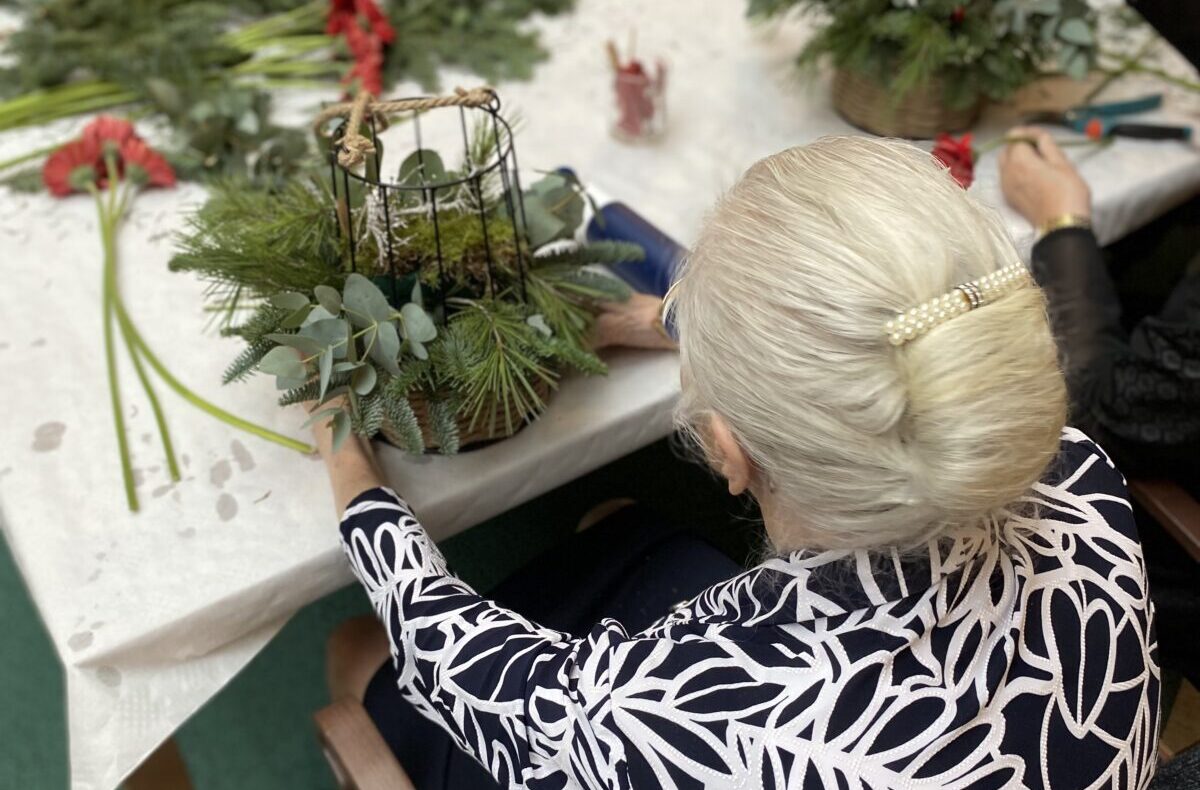 Een oudere vrouw met grijs haar maakt een bloemstuk aan een tafel, omringd door groen en rode bloemen.