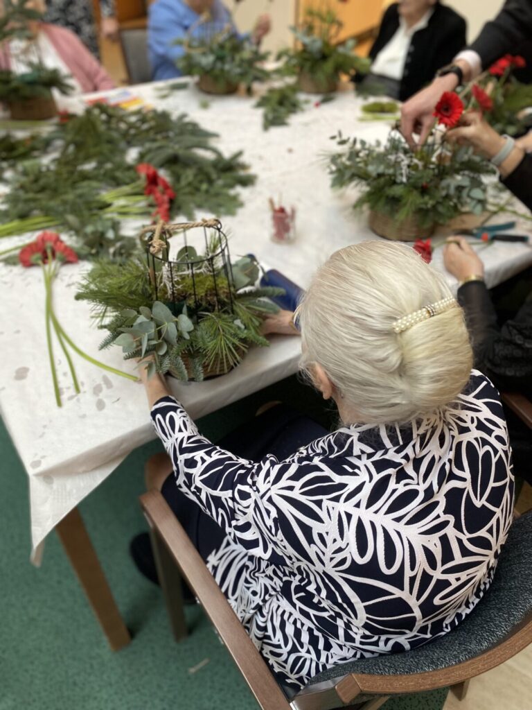 Een oudere vrouw met grijs haar maakt een bloemstuk aan een tafel, omringd door groen en rode bloemen.