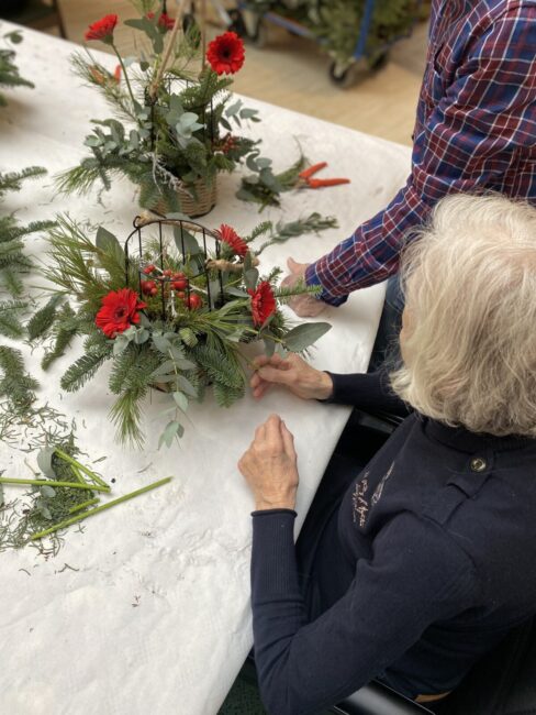 Een oudere vrouw zit aan een tafel en maakt een bloemstuk met rode bloemen en groen blad, geholpen door een andere persoon.