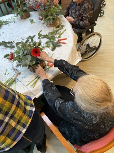 Een oudere vrouw in een rolstoel maakt een bloemstuk met rode bloemen en groen blad aan een ronde tafel met anderen.