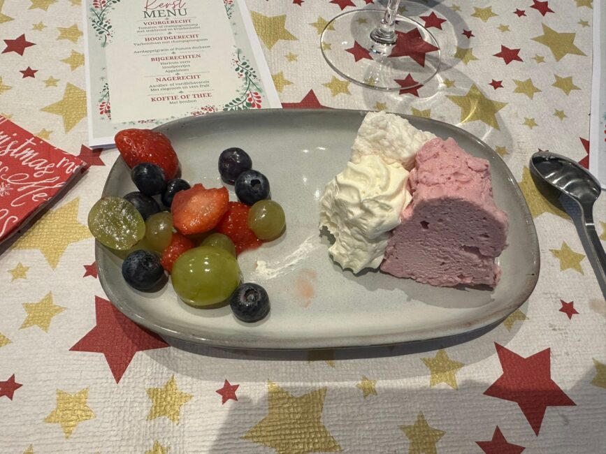 Bord met een dessert van roze mousse, slagroom en vers fruit zoals druiven, aardbeien en bosbessen op een feestelijk tafelkleed.