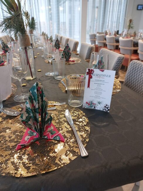 Feestelijk gedekte tafel met gouden placemats, gevouwen servetten en een kerstmenu. Op de achtergrond zijn stoelen zichtbaar.