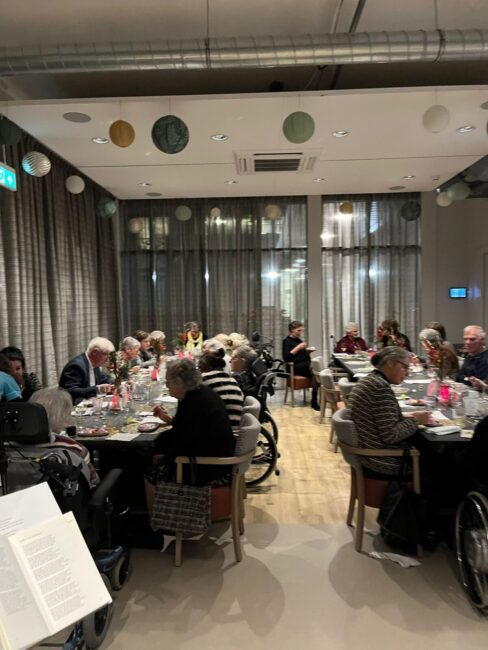 Groep mensen zit aan gedekte tafels in een sfeervol verlichte ruimte voor een kerstdiner. Er hangt een feestelijke sfeer.