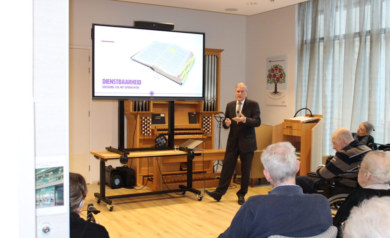 Oudere man in donker pak houdt een presentatie voor een groep oudere mensen.