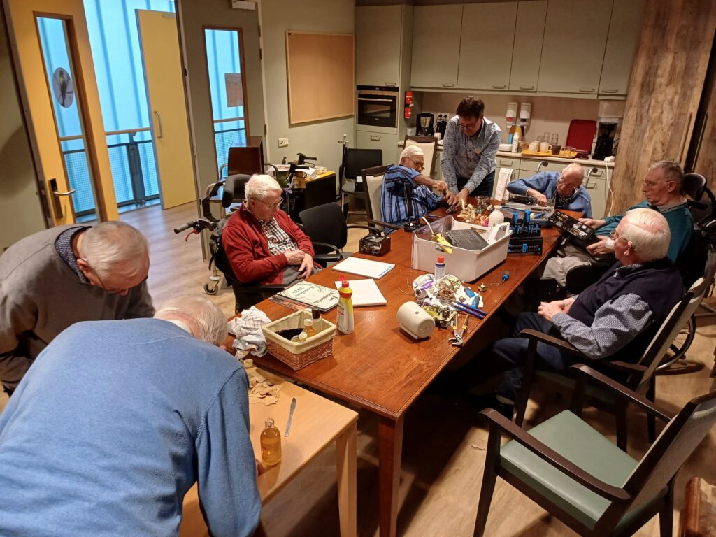 Paar oudere mannen zitten rond een tafel te knutselen met oude apparaten, een man helpt hen daarbij.