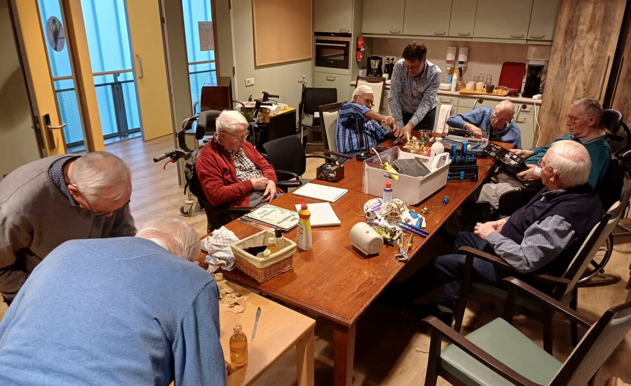 Paar oudere mannen zitten rond een tafel te knutselen met oude apparaten, een man helpt hen daarbij.