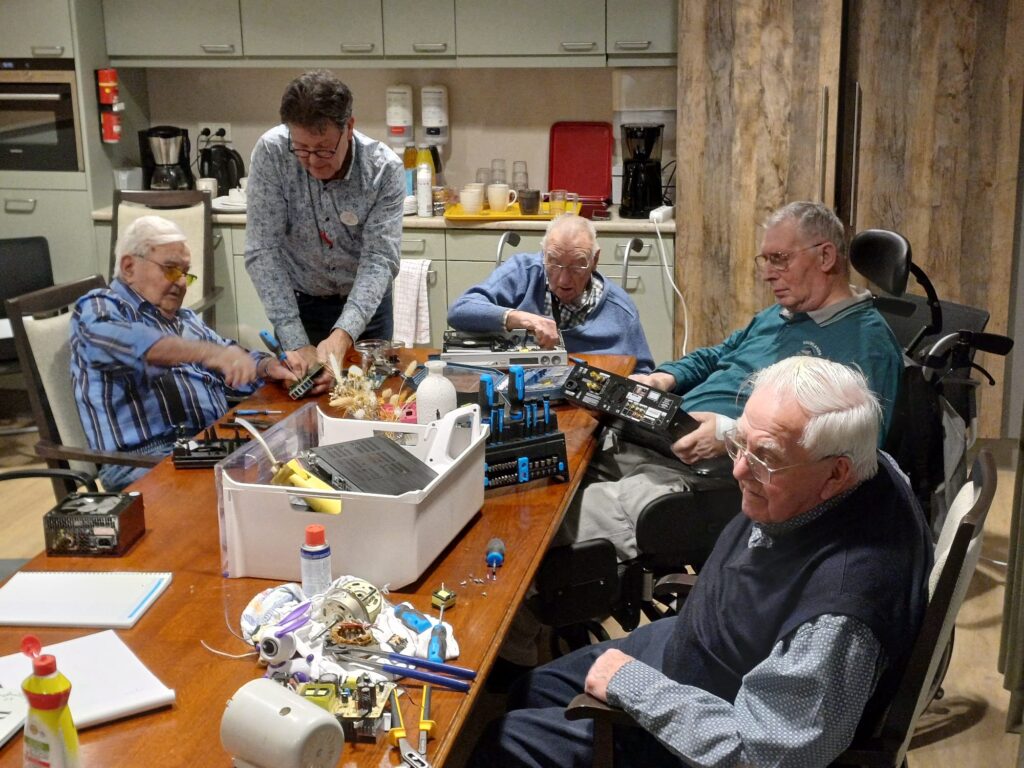 Paar oudere mannen zitten rond een tafel te knutselen met oude apparaten, een man helpt hen daarbij.