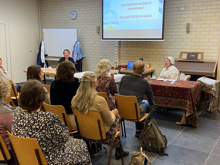 Mensen op stoelen luisteren naar presentatie. Voorin zit een verklede vrouw.