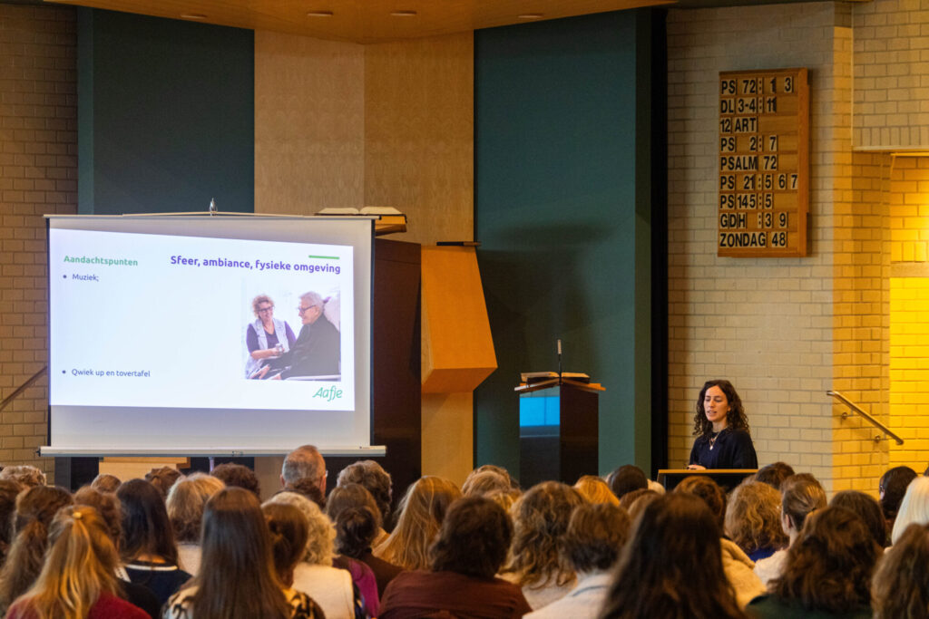Mensen in een kerk luisteren naar de presentatie die een vrouw geeft.