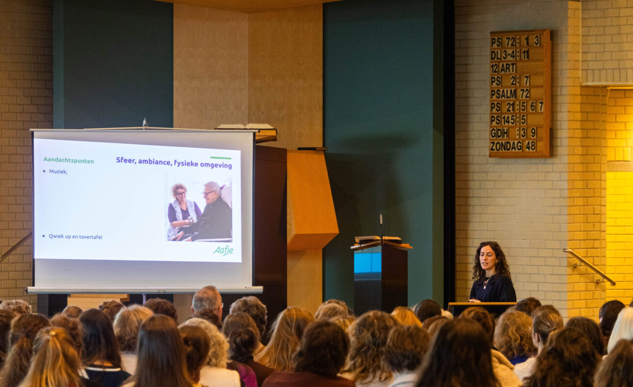 Mensen in een kerk luisteren naar de presentatie die een vrouw geeft.