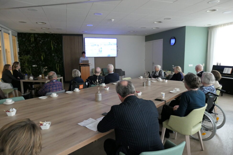 Om een grote vierkante tafel luisteren oudere mensen naar een presentatie die een jongeman houdt.