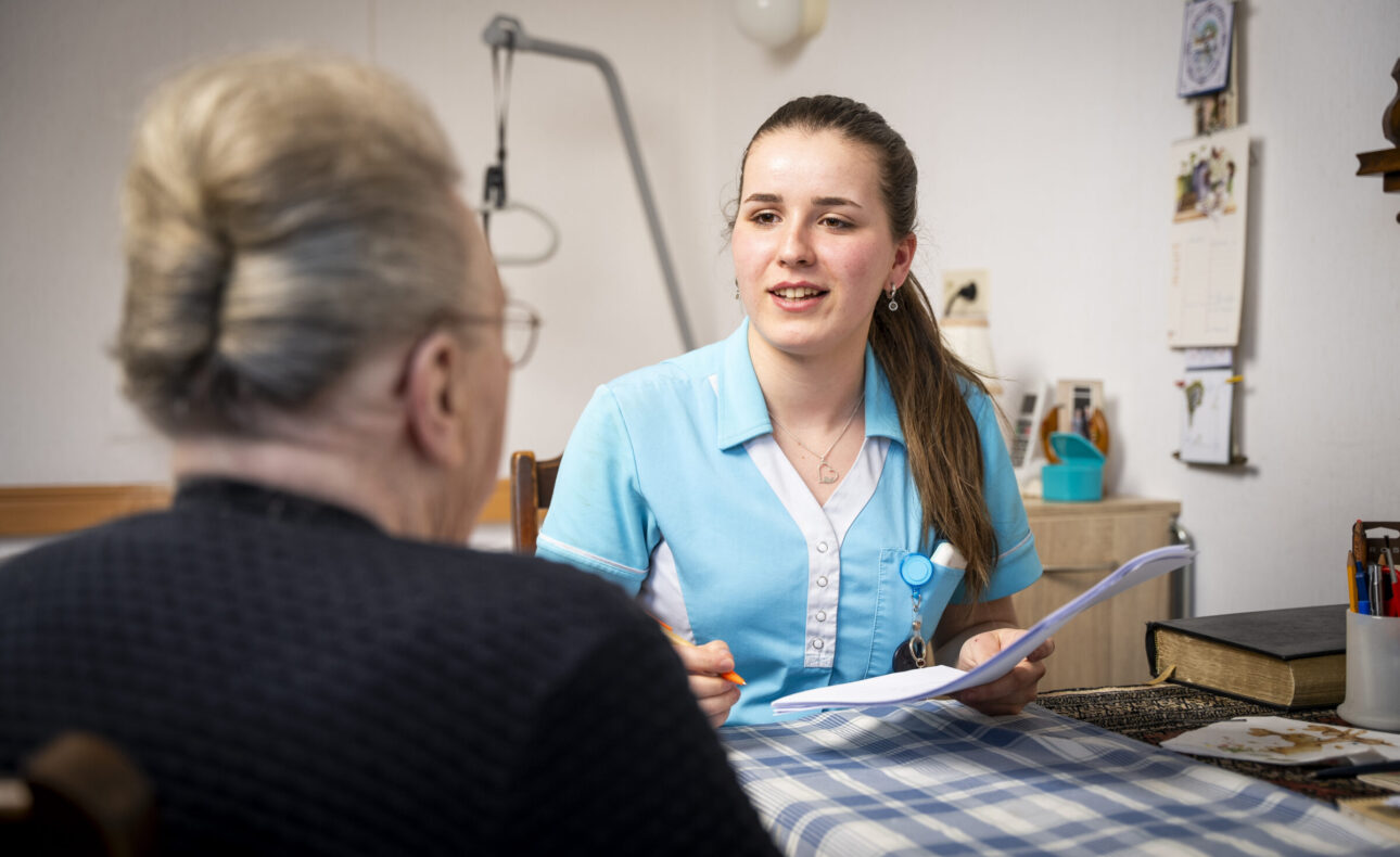 Een zorgverlener in uniform praat met een oudere vrouw aan een tafel, terwijl ze notities maakt op een clipboard.
