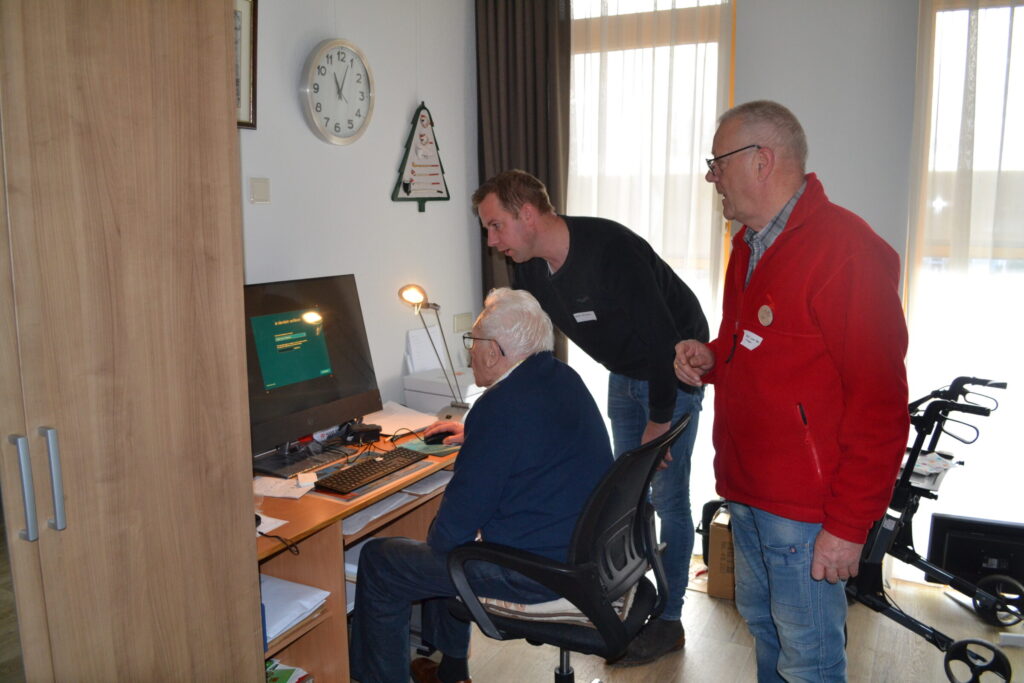 Een oudere man zit achter een computer. Twee jongere kijken met hem mee.
