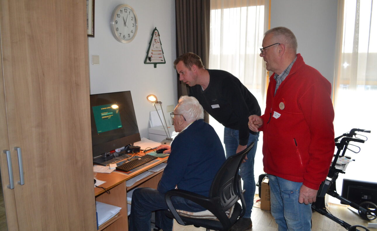 Een oudere man zit achter een computer. Twee jongere kijken met hem mee.