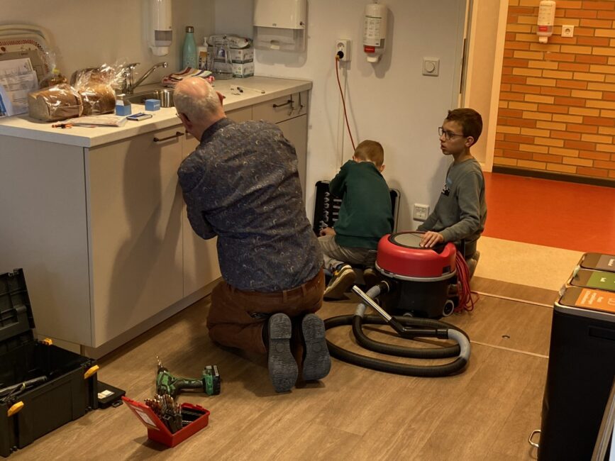 Een man met grijs haar en een blauw overhemd en een bruine broek klust aan een keukenblok. Twee jongens zitten naast hem en helpen hem.