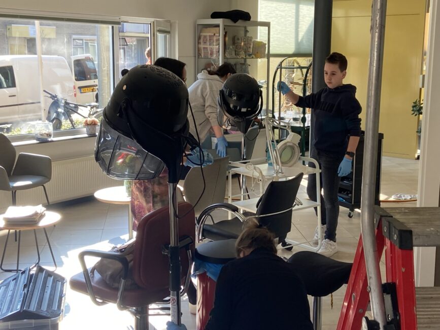 Mensen maken de kapsalon van het woonzorgcentrum schoon. Op de voorgrond een vrouw op de rug, een jongen met een blauwe trui en schoonmaakhandschoenen aan, op de achtergrond nog twee vrouwen met choonmaakhandschoenen aan.