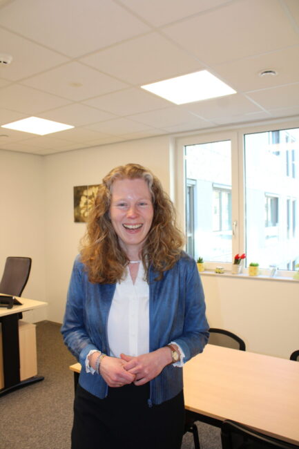 Een vrouw met krullen tot over haar schouders, een blauw vest, witte blouse en donkerblauwe rok kijkt lachend.