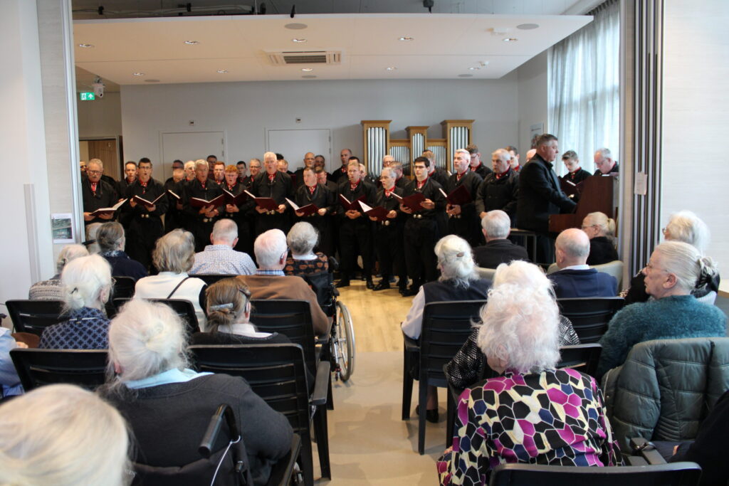 Een mannenkoor in klederdracht is aan het zingen voor een zaal met ouderen.