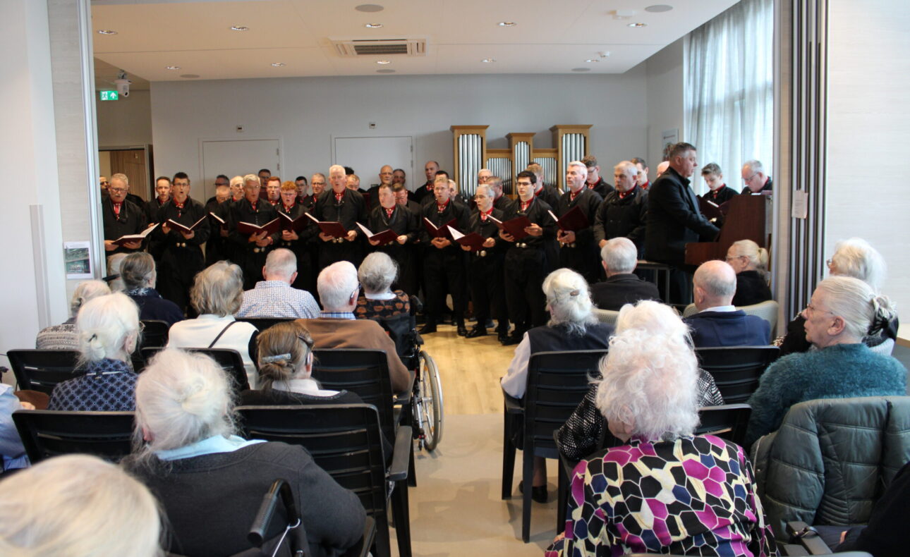 Een mannenkoor in klederdracht is aan het zingen voor een zaal met ouderen.