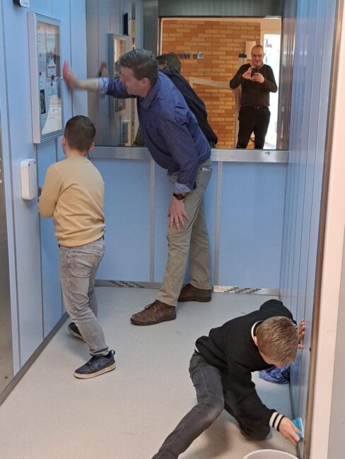 Een man in een blauw overhemd en twee jongens met crème en een zwarte trui maken met een spons de lift schoon.