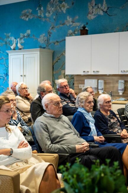 Ouderen in een zaal zitten te luisteren.