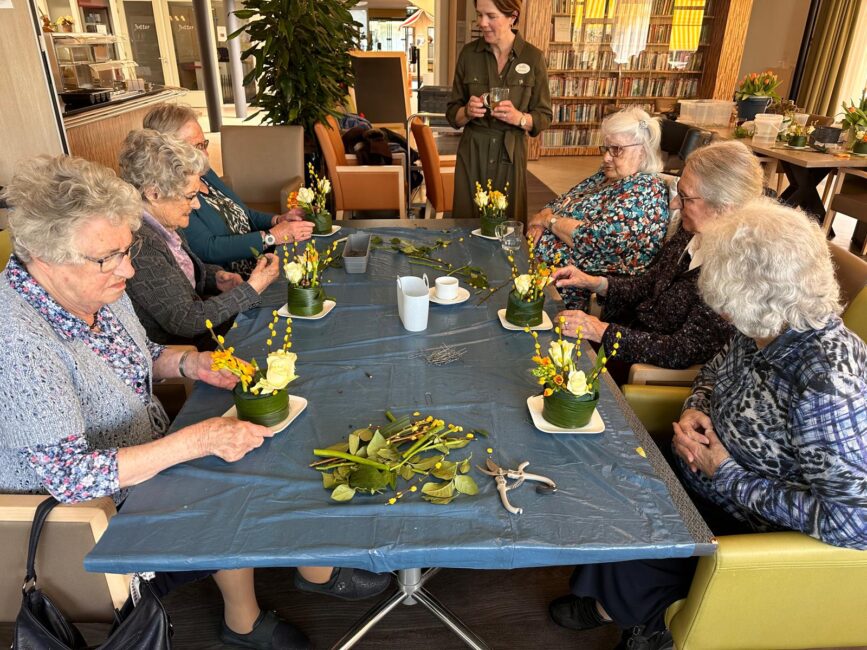 Oudere mensen zitten om een tafel en maken een voorjaarsstukje. Op de achtergrond kijkt een jongere vrouw mee.