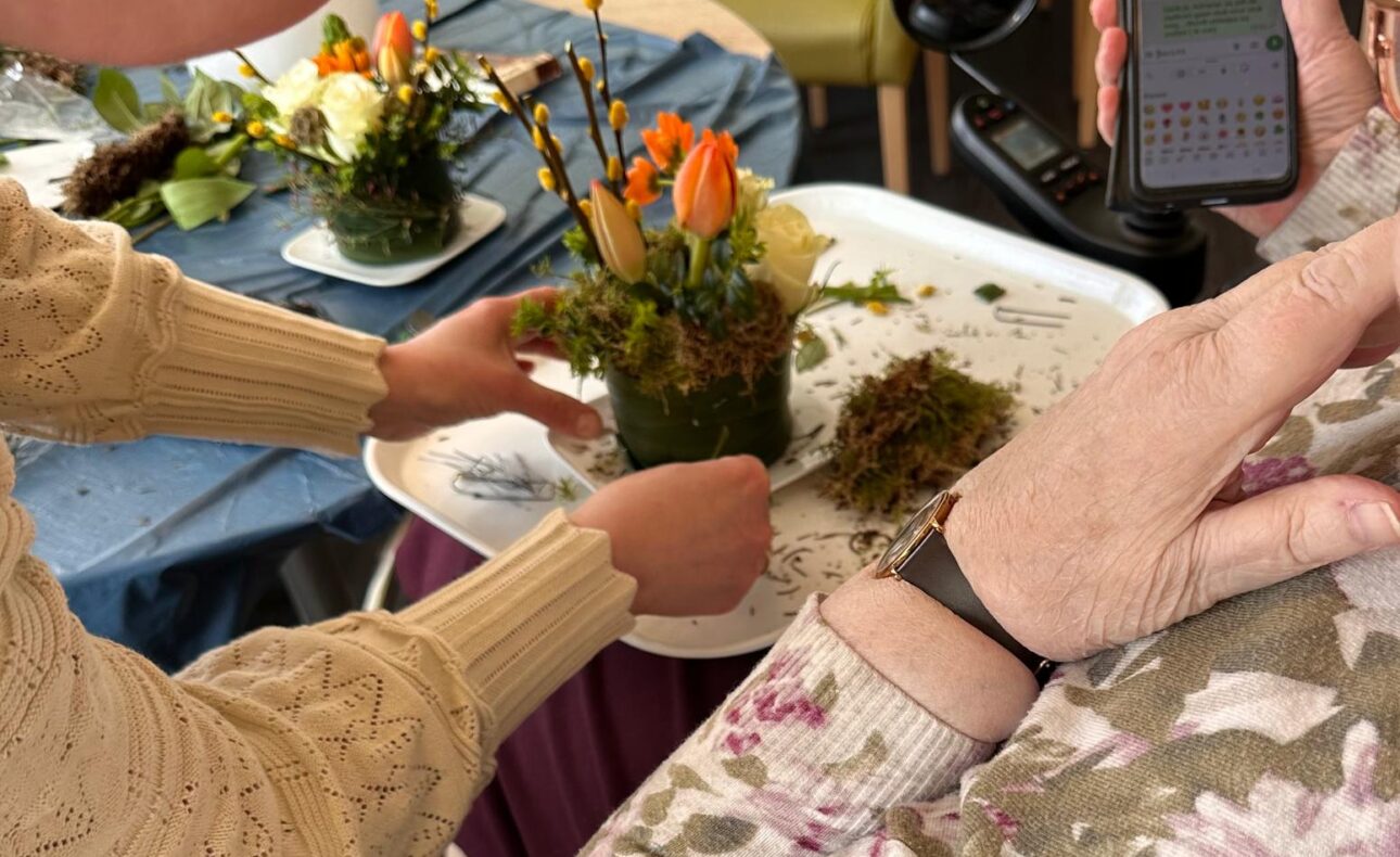 Een jonge vrouw helpt een oudere dame met het maken van een bloemstukje. De oudere dame zit met haar mobiel in haar hand en heeft Whatsapp open staan.