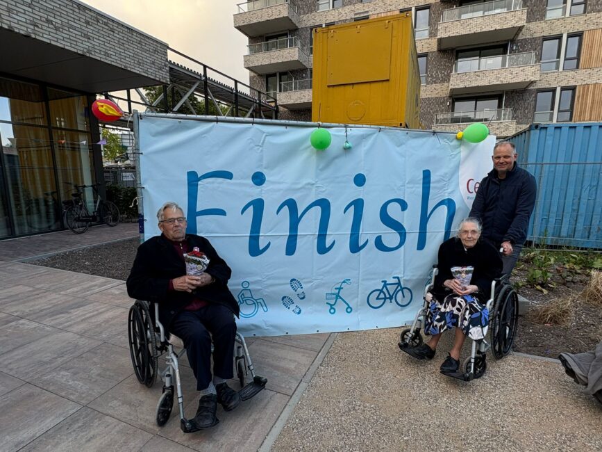 Twee ouderen in rolstoelen staan voor een spandoek met 'finish'. Achter een rolstoel staan een man. Beide ouderen houden een plantje vast.