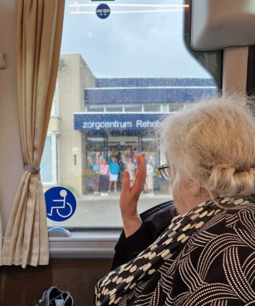 Een oudere vrouw in een bus zwaait naar mensen voor de ingang van Zorgcentrum Rehoboth.