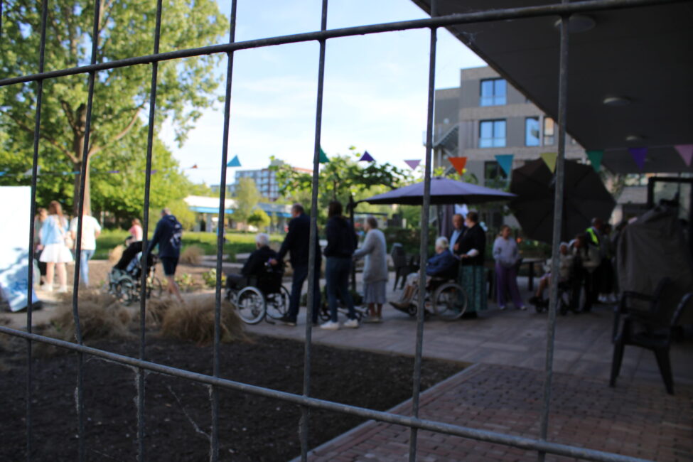 Door de spijlen van een hek zien we ouderen in rolstoelen en mensen daarbij op een terras.