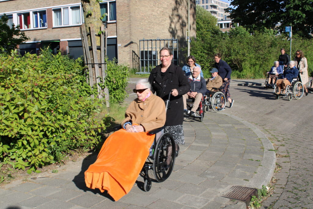 Een vrouw in een rolstoel met een deken over haar benen wordt geduwd door een vrouw. Op de achtergrond zijn meerdere ouderen te zien die in rolstoelen worden geduwd.