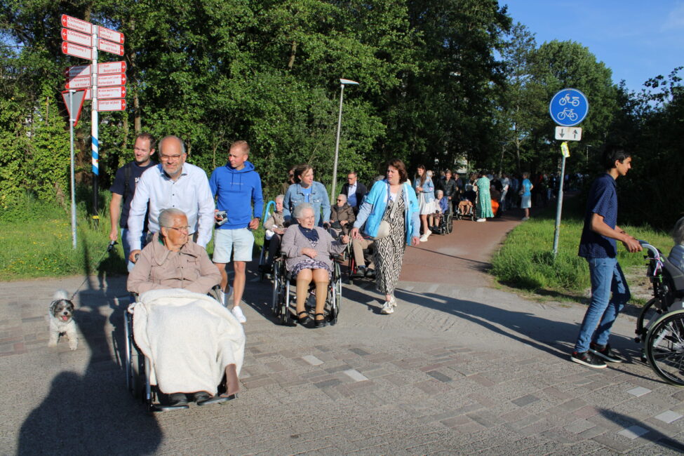 Een groep ouderen in rolstoelen worden over een fietspad geduwd. Mensen lopen mee.