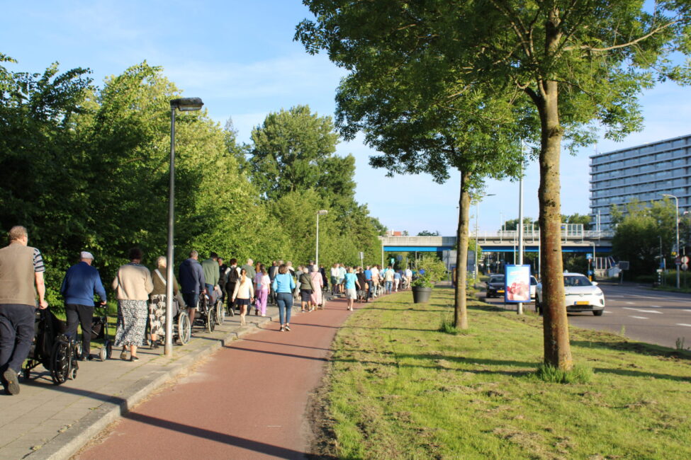 Een groep ouderen in rolstoelen op een stoep met mensen die duwen en meelopen. Naast de stoep is een fietspad.