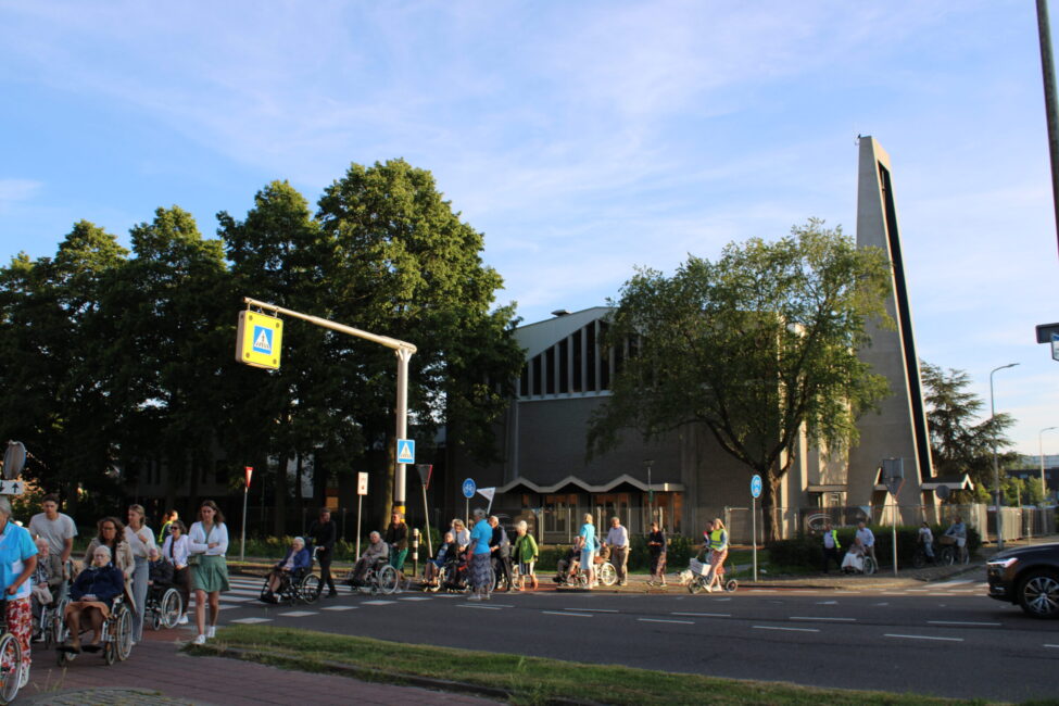 Een groep ouderen in rolstoelen die worden geduwd steken een weg over. Op de achtergrond is een kerk te zien.