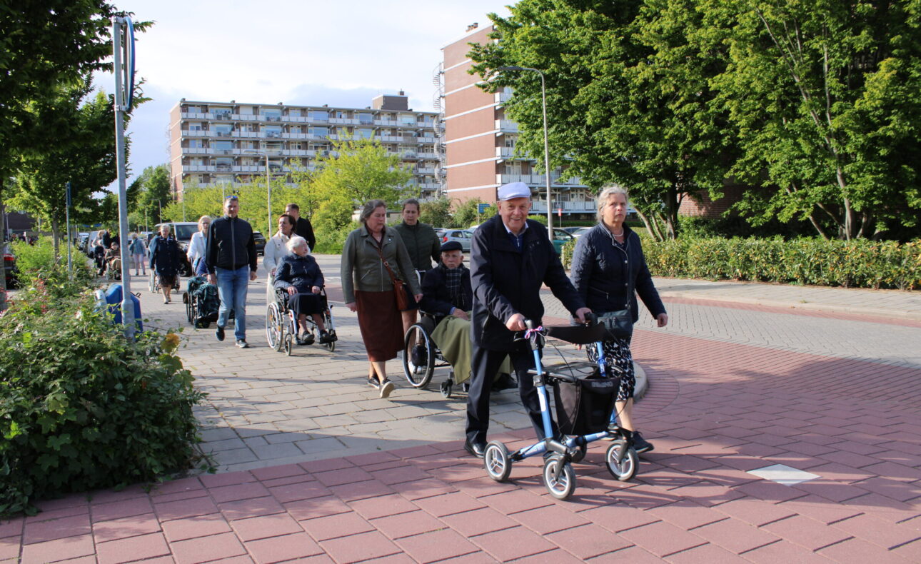 Een man loopt achter een rollator, naast hem loopt een vrouw. Daarachter zijn meerdere rolstoelen te zien met mensen die meelopen.