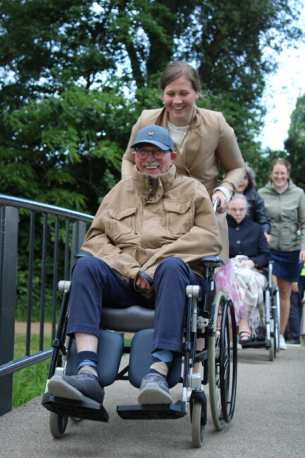 Een man in een rolstoel wordt geduwd door een vrouw over een brug. Op de achtergrond is nog een rolstoel te zien.