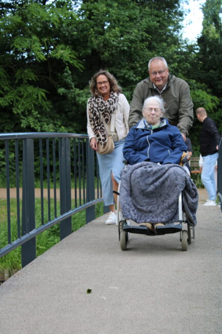 Een vrouw in een rolstoel wordt geduwd door een man. Op de achtergrond loopt nog een vrouw. Ze lopen over een brug.