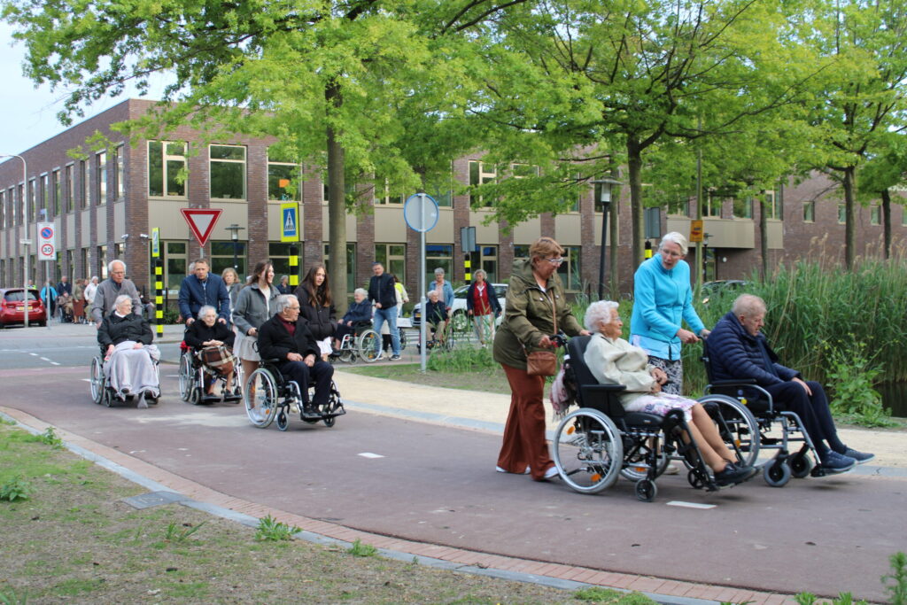 Ouderen in rolstoelen worden geduwd over een fietspad. Op de achtergrond een gebouw.