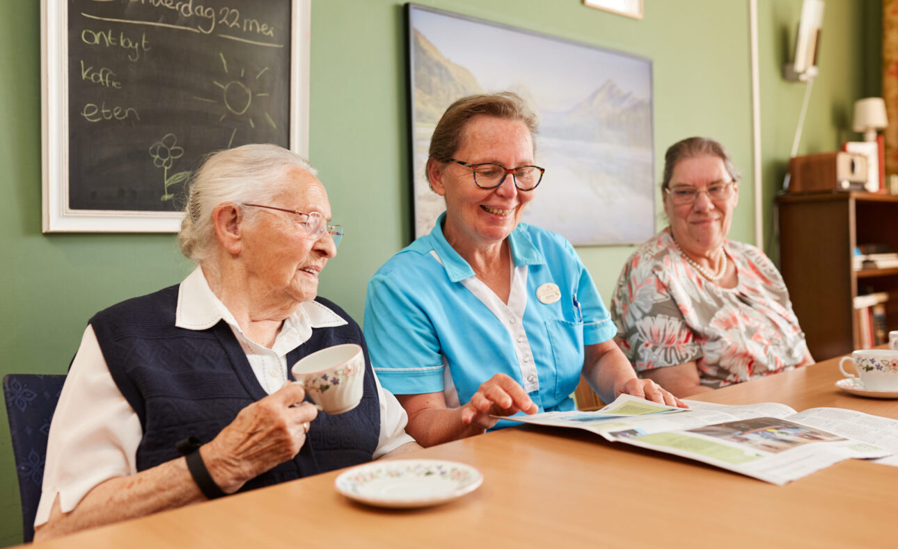 Zorgmedewerker die met twee ouderen de krant leest aan tafel.