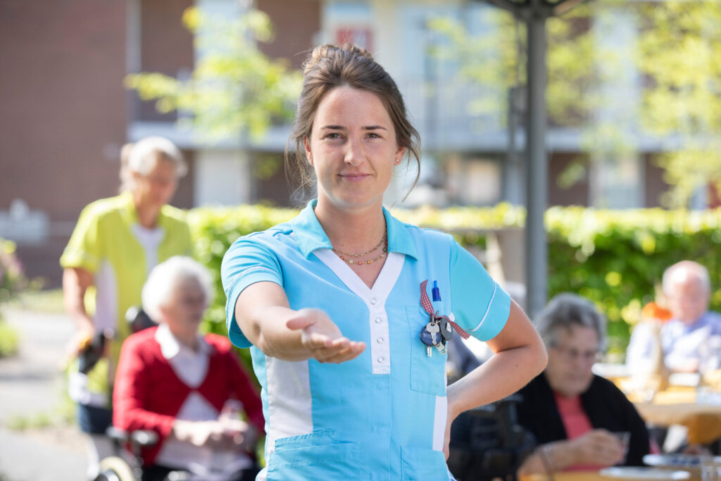 Een jonge vrouw in een blauw Cedrah-jasje houdt haar hand op. Op de achtergrond zijn vaag een paar ouderen te zien.