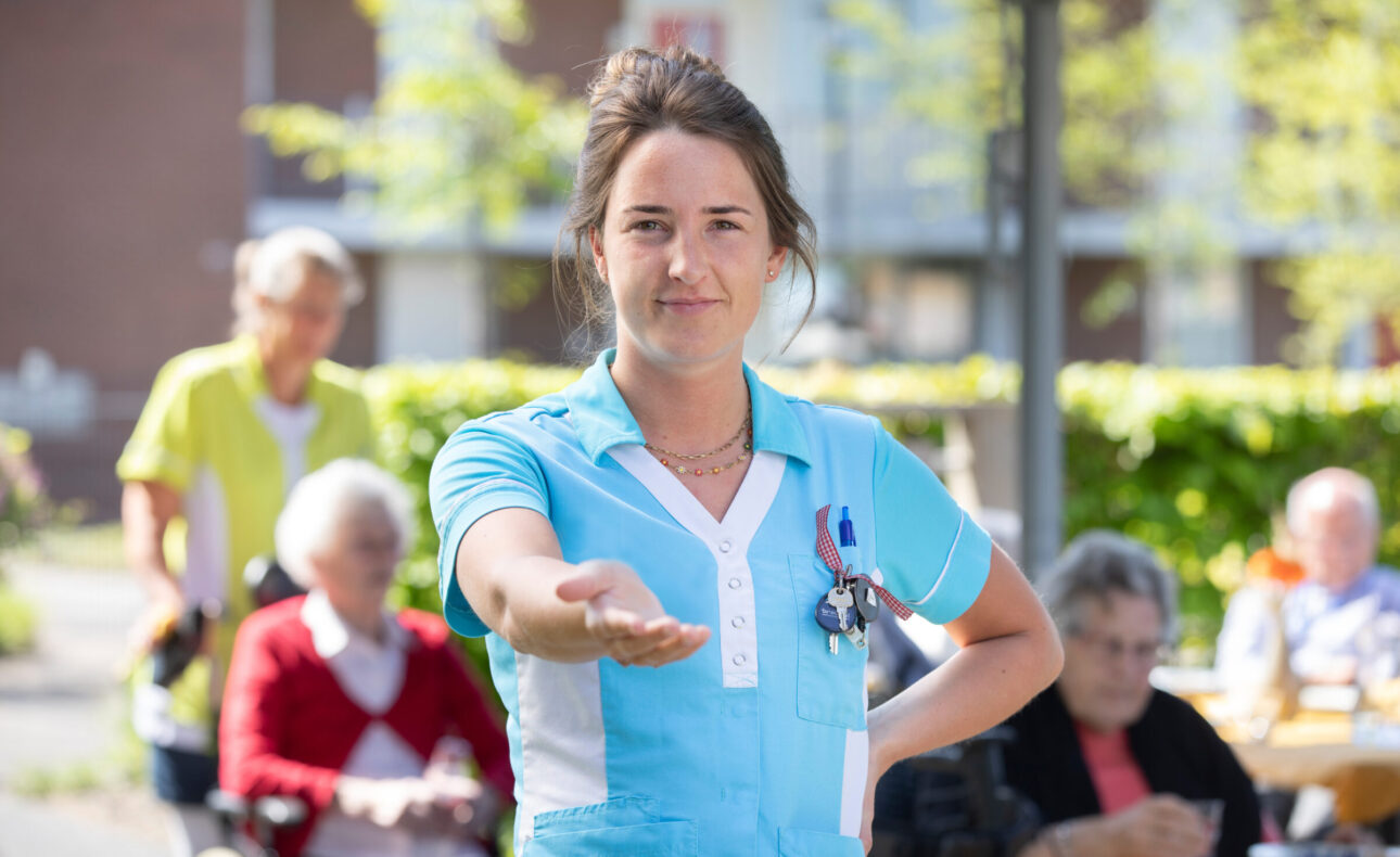 Een jonge vrouw in een blauw Cedrah-jasje houdt haar hand op. Op de achtergrond zijn vaag een paar ouderen te zien.