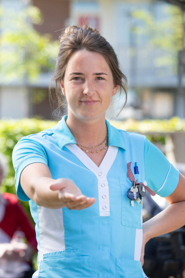 Een jonge vrouw in een blauw Cedrah-jasje houdt haar hand op. Op de achtergrond zijn vaag een paar ouderen te zien.