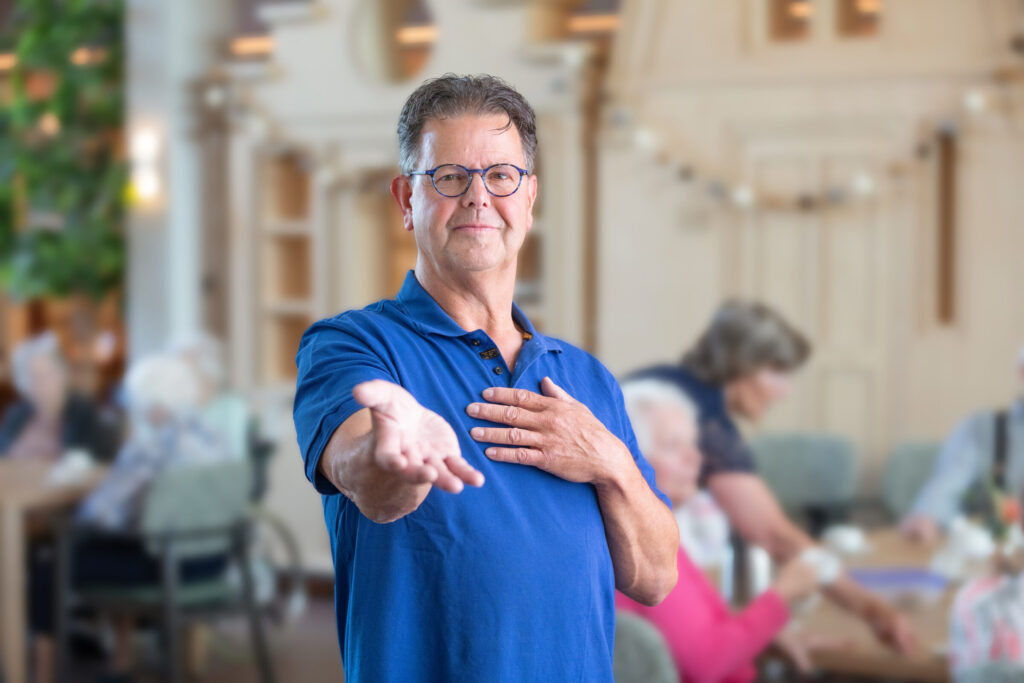 Een man in een blauw shirt houdt zijn hand op. Op de achtergrond zijn vraag enkele ouderen te zien.