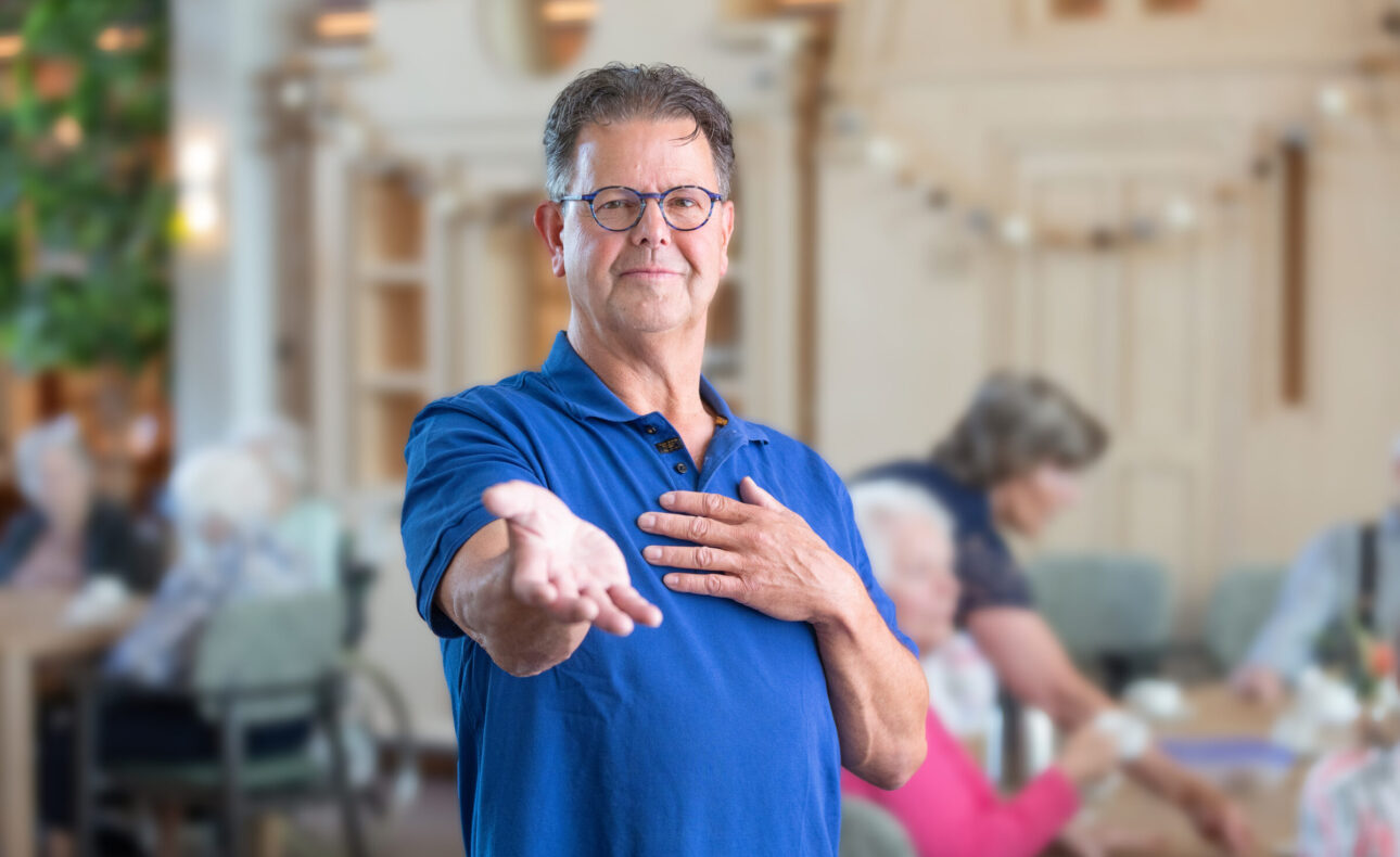 Een man in een blauw shirt houdt zijn hand op. Op de achtergrond zijn vraag enkele ouderen te zien.