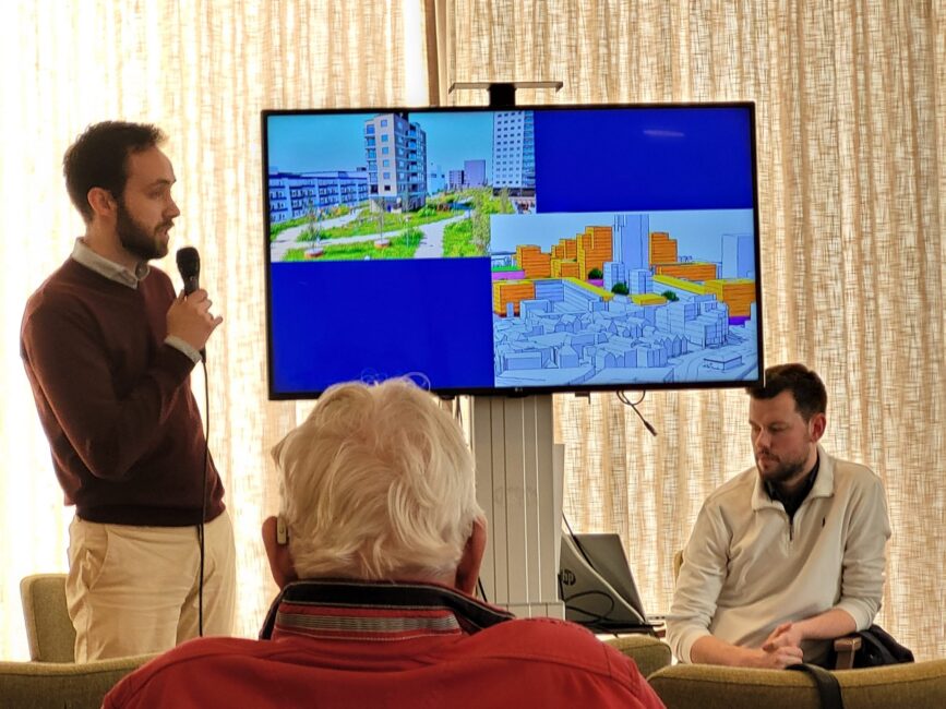 Man met microfoon geeft presentatie. Op de achtergrond een scherm met gebouwen.