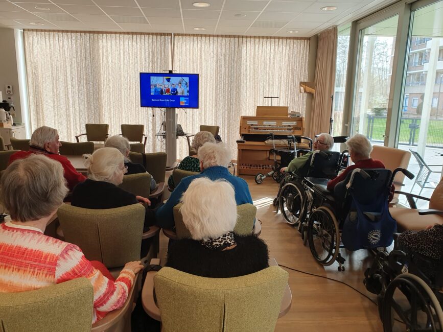 Ouderen in een zaal kijken naar een presentatie op een scherm.